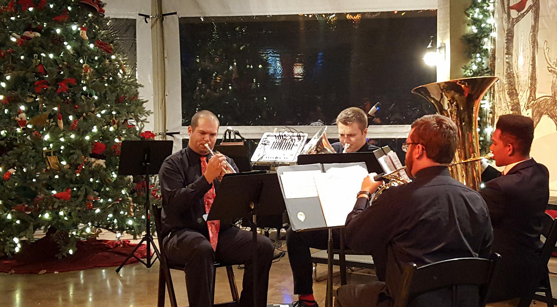 Four individuals play brass instruments in a room decorated for Christmas